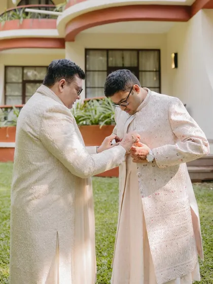 A touching moment as the groom gets help with his sherwani from a family member in a sunlit garden.