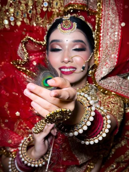 A creative shot with a peacock feather, adding a touch of traditional artistry. The bride's red and gold outfit is complemented by her classic makeup and joyful expression.