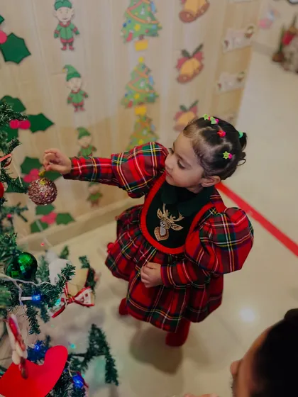 A close-up of a little girl in a festive plaid dress carefully placing an ornament on the tree. These moments of focused participation are what make the holidays special.