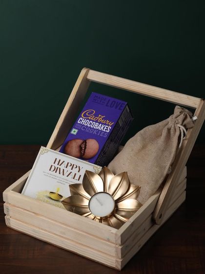 This simple and sweet hamper in a wooden basket is a perfect budget-friendly option. It includes Cadbury Chocobakes, a golden flower diya, and a potli of goodies.
