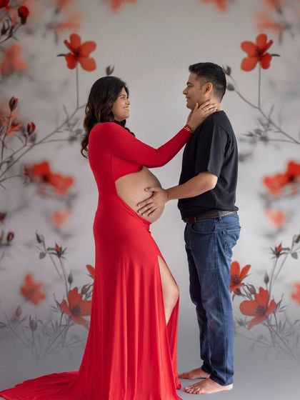 A tender moment from a studio session with a floral backdrop. The mom-to-be in a striking red dress gently touches her partner's face.