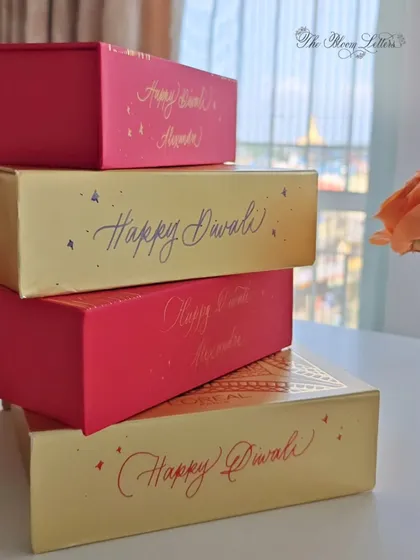 A stack of festive gift boxes personalized for Diwali. The combination of red and gold boxes with calligraphy in different colors creates a vibrant and joyful presentation for holiday gifting.