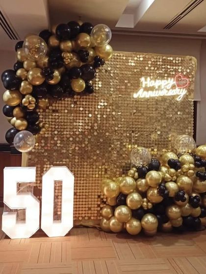 Celebrating a golden anniversary with a spectacular gold sequin backdrop. This decoration for a 50th anniversary features marquee numbers, a "Happy Anniversary" neon sign, and a lush garland of black and gold balloons.