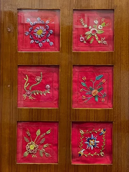 A set of six small, framed floral embroideries on a vibrant red background. These pieces are available for purchase and would look lovely as a collection in any room.