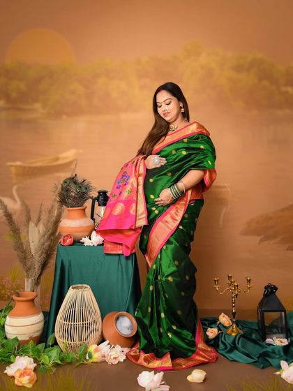 A graceful solo portrait of the mom-to-be in a dark green and pink Paithani saree. The pose highlights the beautiful drape of the saree and the baby bump against our traditional painted backdrop.