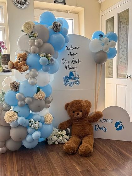 A sweet 'Welcome Home Our Little Prince' setup. This decoration includes a custom backdrop, balloon arrangements in blue, grey, and white, and adorable teddy bears to welcome the newborn.