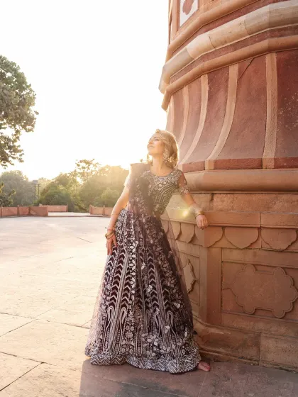 A full-length shot of the model leaning against a historic sandstone pillar. The warm sunlight and her upward gaze create a feeling of serenity and grace, showcasing the full splendor of the lehenga.