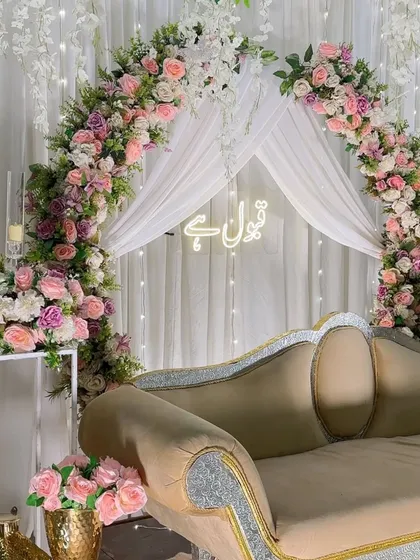 An elegant and serene Nikah backdrop. This setup features a beautiful floral arch in shades of pink and white, with delicate white drapes and a glowing 'Qubool Hai' neon sign, creating the perfect atmosphere for the ceremony.