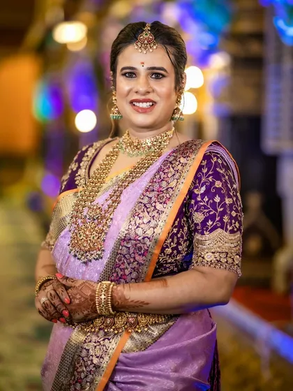 A close-up portrait showcasing the detailed hand embroidery on the purple blouse. The gold threadwork and patterns are designed to complement the client's traditional gold jewelry.