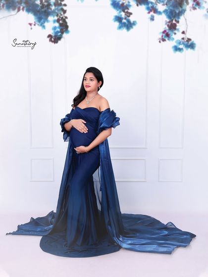 A full-length portrait of a mother-to-be in a deep blue gown. The simple background and elegant pose highlight the beauty of the dress and her glowing silhouette.
