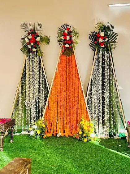 This is a modern and minimalist Haldi backdrop we created. The design uses three triangular frames with lush green turf as a base, featuring vibrant orange marigold curtains and white floral strings, topped with fresh flower bouquets for a clean and contemporary look.