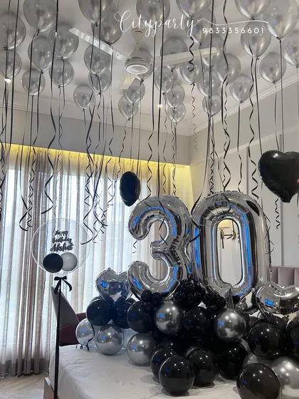 A chic and modern 30th birthday surprise decoration using a black and silver color scheme. The setup includes large silver '30' foil balloons, a bed arrangement, and matching balloons on the ceiling.