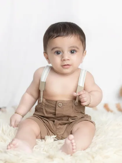 A cute baby boy in suspenders, sitting confidently for his portrait.