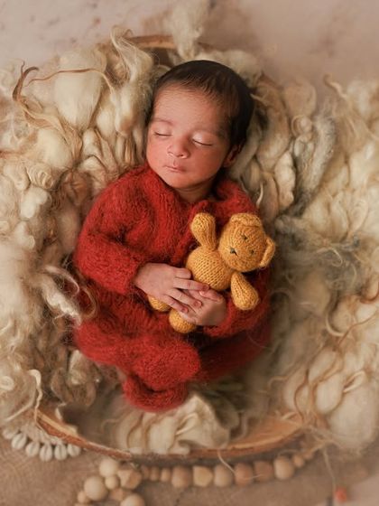 Hello, tiny miracle. This baby looks so cozy in a vibrant red knit romper, snuggling a little teddy bear in a nest of soft wool.