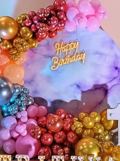 A colorful and modern first birthday setup. The marbled backdrop is framed by a rainbow of metallic balloons, with the name and number in marquee lights.