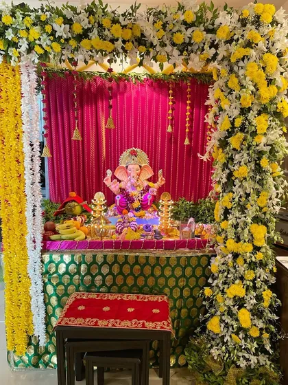 A bright and cheerful Ganpati setup featuring a full arch of yellow and white flowers against a vibrant pink backdrop. Hanging bells add a traditional touch to this sunny design.