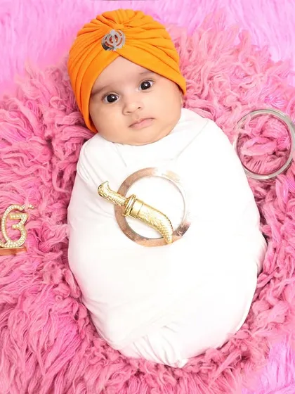 A full-body shot of a newborn swaddled in white for his Sikh-themed session. The orange turban provides a beautiful pop of color, and the props are arranged artfully on a plush pink rug.
