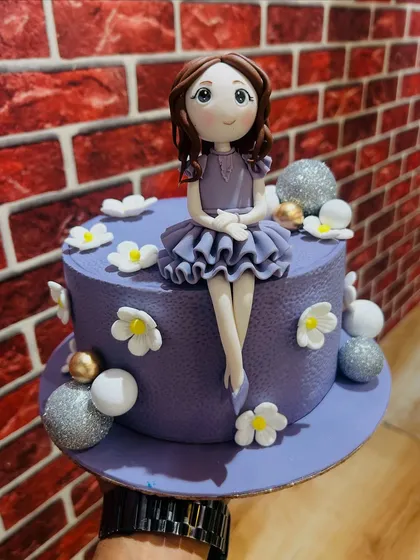 A lovely purple cake with a sitting doll figurine, surrounded by delicate white flowers.