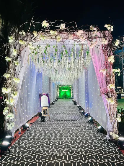 This magical Sangeet entrance features a rustic branch archway and a tunnel of hanging white wisteria and fairy lights, creating a path that feels like walking through an enchanted forest.