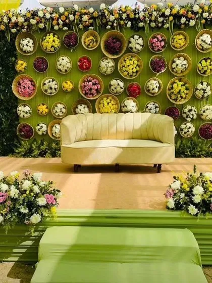 A beautiful Mehendi seating area with a backdrop made of a green wall and colorful flower baskets. The off-white sofa provides a comfortable and stylish spot for the bride.