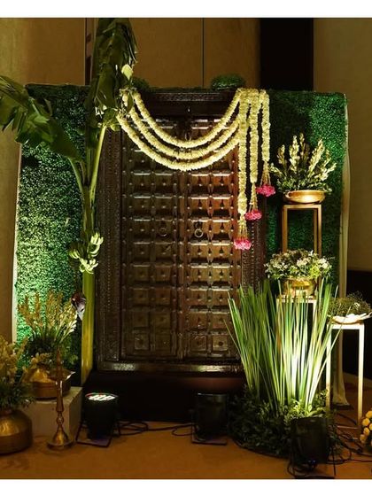 A rustic and traditional photo corner featuring an old wooden door, adorned with jasmine garlands and surrounded by banana plants and brass pots.