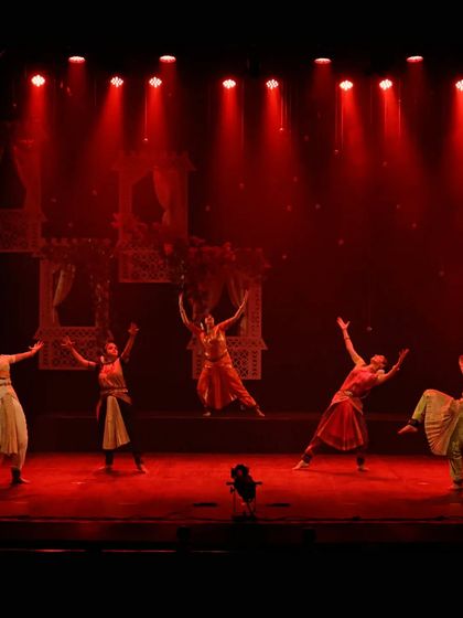 A wide shot of the ensemble on stage during "UNMASKED". The red lighting and expansive poses create a scene of high drama and energy, showcasing the scale of the production.