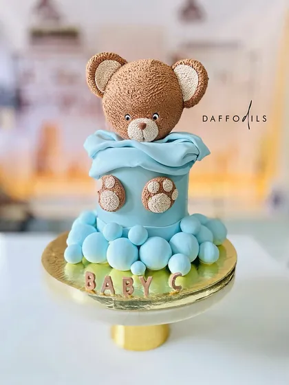 A cute and cuddly teddy bear cake for Baby C. The cake features a sculpted teddy peeking out of a blue blanket, surrounded by fluffy bubbles.