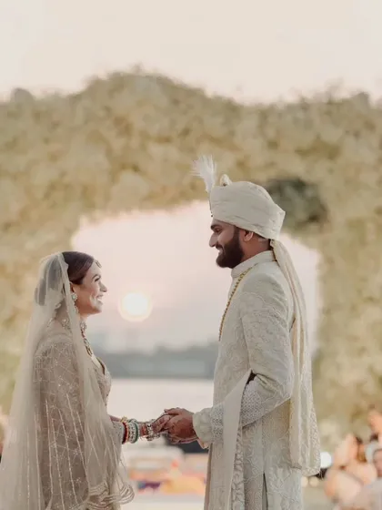 A quiet moment of connection between the bride and groom, holding hands before their floral arch. The setting sun paints the sky, creating a serene and romantic atmosphere for their sunset wedding.