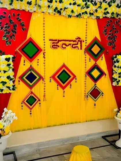 A bold and bright Haldi backdrop using a combination of red and yellow fabrics. The look is completed with colorful geometric hangings and marigold flower details, perfect for a lively and energetic celebration.