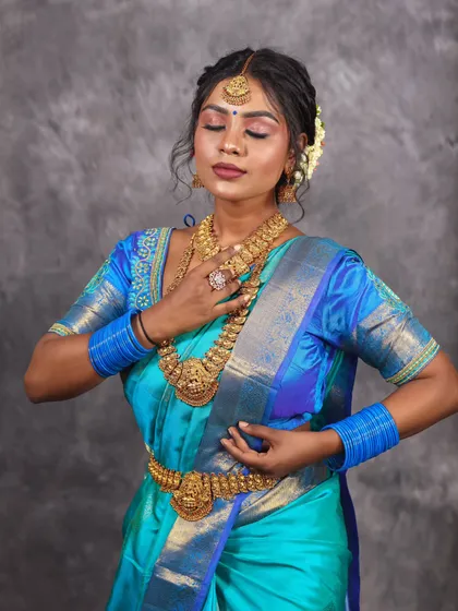 A graceful pose in a blue Kanchipuram saree, showing the intricate gold jewelry and the traditional floral hair arrangement. The studio backdrop keeps the focus entirely on the ethnic ensemble.