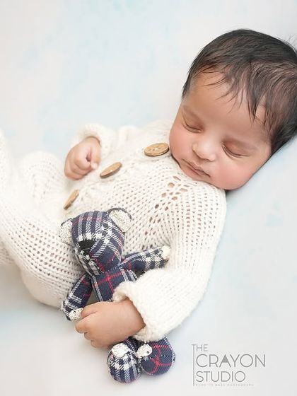 A newborn in a classic white knit outfit cuddles a tiny plaid teddy bear, creating a timeless and heartwarming portrait against a soft blue background.