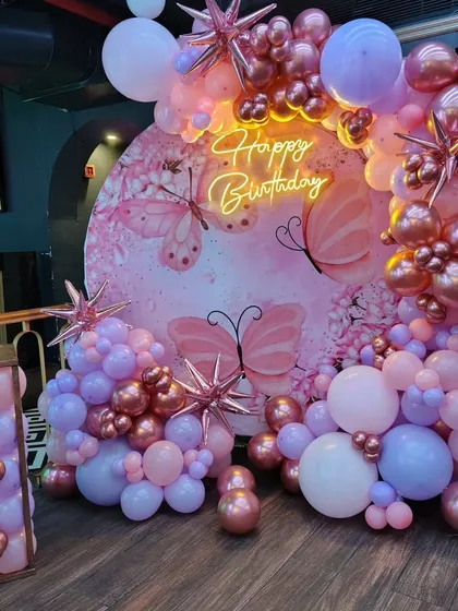 A butterfly-themed birthday decoration in shades of pink, purple, and rose gold. This setup includes a custom printed backdrop, a full balloon garland, and star-shaped foil balloons.
