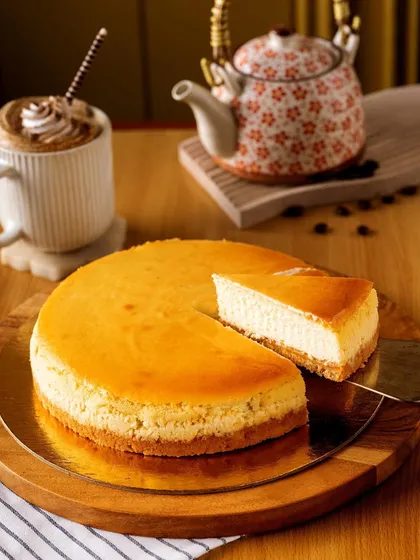 A classic New York cheesecake, with one slice removed to invite you in. The simple wooden board and accompanying coffee create a relaxed, cafe-style atmosphere, making the dessert feel accessible and delicious.