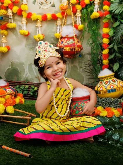 A joyful smile from a little girl dressed as Radha, holding a pot in the colorful Janmashtami scene.
