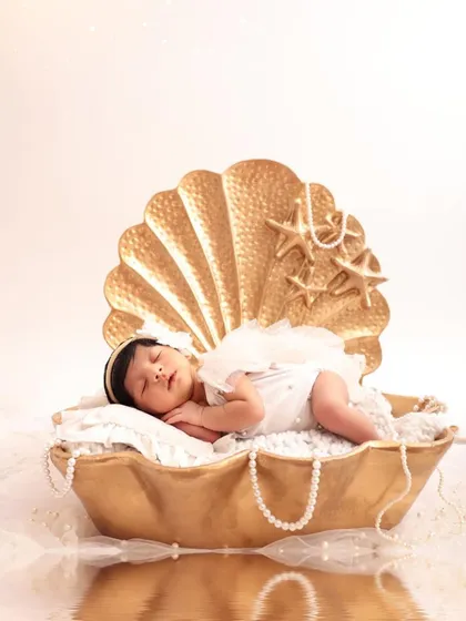 A newborn baby, adorned with angel wings, sleeps peacefully in a golden seashell prop. This portrait is part of a sibling shoot, symbolizing the new baby as a precious pearl.