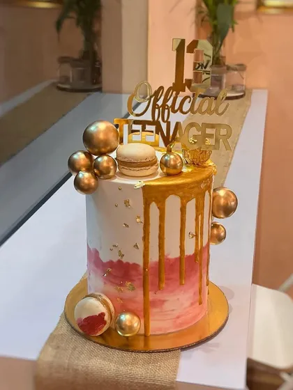 A beautiful cake to welcome a new teenager. This design features a pink and white watercolor effect, a rich gold drip, and is decorated with golden spheres and macarons for a trendy and celebratory look.