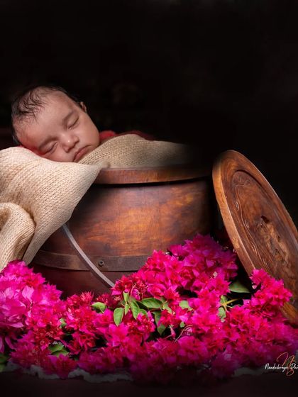 For a touch of rustic charm, I often use natural props like this wooden bucket. The contrast between the dark wood, the bright bougainvillea flowers, and the baby's soft skin makes for a stunning and unique photograph.