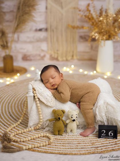 A cozy, bohemian-inspired setup with a baby in a brown knit outfit sleeping on a miniature bed with fairy lights twinkling in the background.