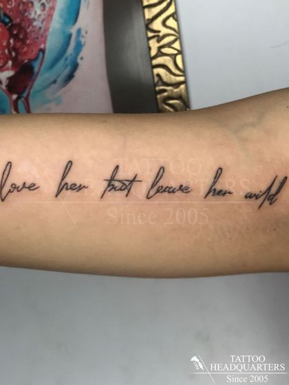 The quote "love her but leave her wild" in a delicate, handwritten script along the forearm, a popular choice for expressive text tattoos.
