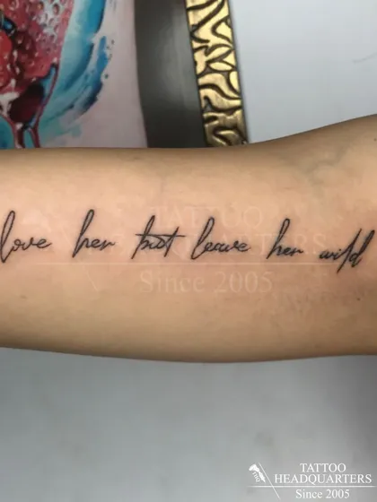The quote "love her but leave her wild" in a delicate, handwritten script along the forearm, a popular choice for expressive text tattoos.