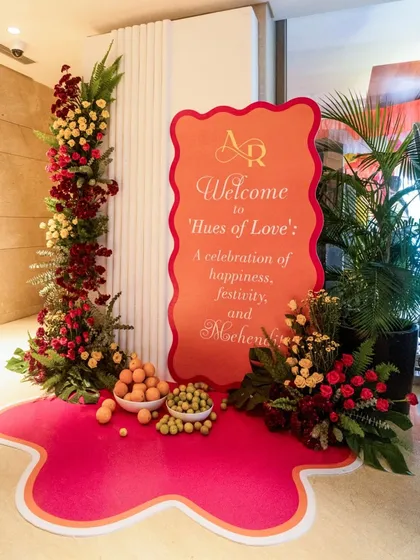 The welcome signage for the "Hues of Love" Mehendi celebration. The entrance is decorated with a vibrant pink floor decal, columns of red and yellow flowers, and bowls of fresh fruit.