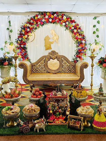 An elegant baby shower setup with a floral arch and an ornate sofa. The stage is decorated with traditional dolls, brass lamps, and a beautiful arrangement of offerings on a green turf.