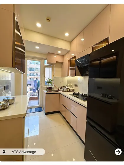 A beautiful and functional L-shaped modular kitchen in a soft pink hue. The design incorporates glossy cabinets, a white countertop, and a black chimney and fridge for a modern, contrasting look.