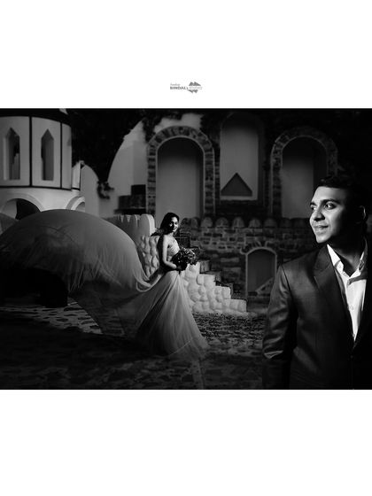 A cinematic black and white shot where the bride's flowing gown takes center stage. The groom looks on, creating a narrative of admiration and romance.
