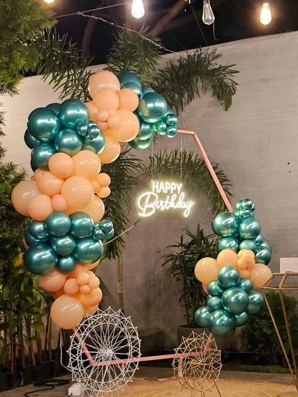A modern and artistic birthday decoration. This setup features a unique balloon arrangement in teal and peach on a pink frame, complemented by a "Happy Birthday" neon sign and a decorative bicycle prop.