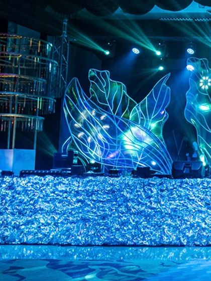 A close-up of the Sangeet stage decor, featuring the shimmering silver facade and the large, illuminated leaf-like structures. The cool blue lighting gives the entire setup a futuristic and glamorous feel.