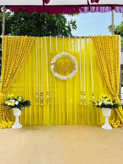 An elegant and bright Haldi backdrop created with yellow drapes, strings of white rajnigandha flowers, and a central floral wreath. The marigold borders and flower pots complete this classic look.