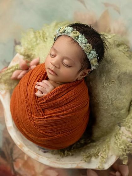 A beautiful composition featuring a baby in a rust colored wrap, resting in a prop against a painterly floral backdrop. The colors and textures work together to create a true work of art.