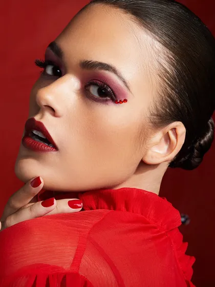 A classic red-on-red look with a bold red lip, red eyeshadow, and delicate red rhinestone details in the inner corner of the eye.
