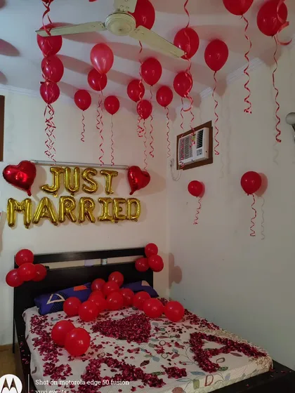 An all-red theme for a first night room decoration. This setup includes red balloons on the ceiling, 'Just Married' foil letters, and rose petals with the couple's initials on the bed.